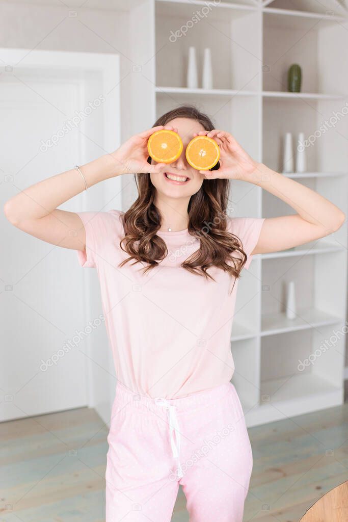 Young woman in pink house pajamas holding sliced oranges at the eyes on light background