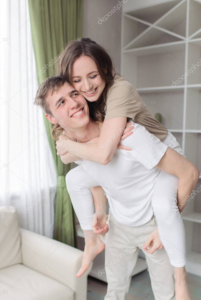 Cute cheerful couple smiling toothily with girl riding man back - piggyback - at home. Cozy apartment of a young family.
