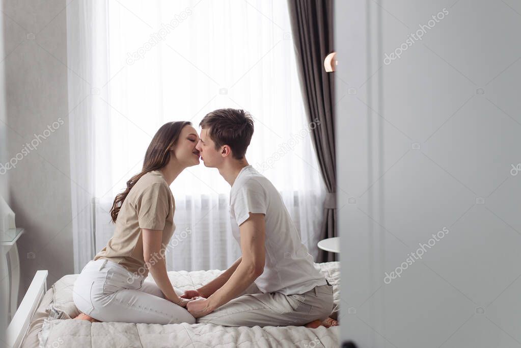 A young couple of lovers sit and lie on a bed in a home bedroom. The guy and the girl hug and kiss. Romantic time together.