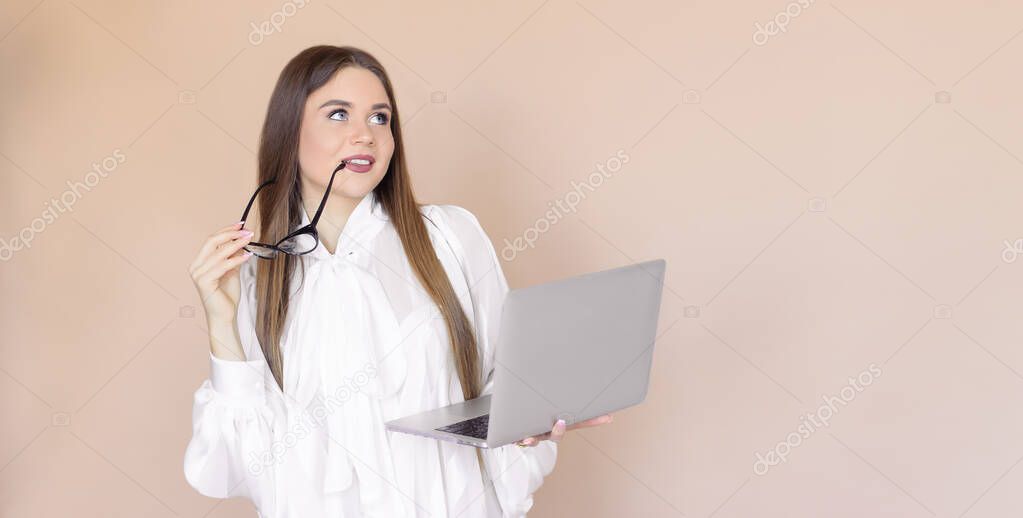 The girl uses a laptop, she 's business, wearing glasses and a white shirt. A young woman with a computer on a beige one-ton background.