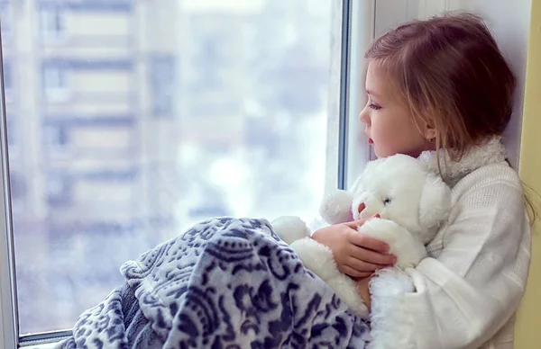 Linda niñita abrazando a un oso de peluche. Un bebé lindo en la habitación se sienta en la ventana en el invierno —  Fotos de Stock