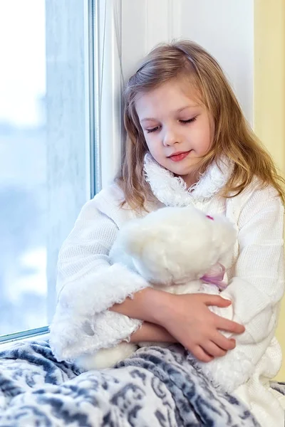 Cute little girl hugging a teddy bear. A cute baby in the room sits at the window in the winter — Stock Photo, Image