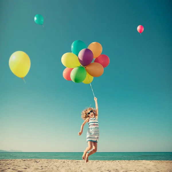 Niño feliz saltando con globos de colores en la playa de arena —  Fotos de Stock