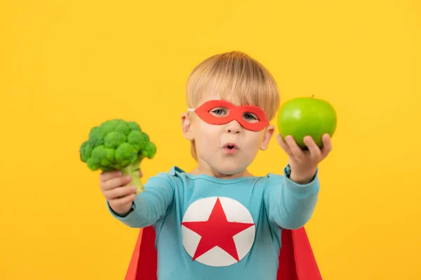 Superheldenkind hält Brokkoli und Apfel in der Hand — Stockfoto