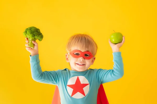Superhjälte barn som håller broccoli och äpple — Stockfoto