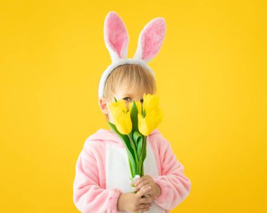 Sarı arka planda Paskalya tavşanı takan komik çocuk. Bir buket çiçek tutan mutlu çocuk. Bahar tatili kavramı