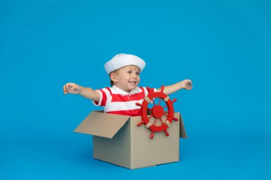 Mutlu çocuk kaptan olmayı hayal ediyor. Çocuk mavi arka planda eğleniyor. Çizgili tişörtlü çocuk karton kutuda oynuyor. Yaz tatili ve seyahat konsepti. Hayal ve hayal gücü