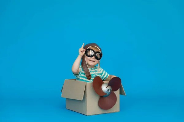 Happy child dreams of becoming a pilot. Kid having fun against blue paper background. Boy wearing striped shirt playing in cardboard box. Summer vacation and travel concept. Dream and imagination