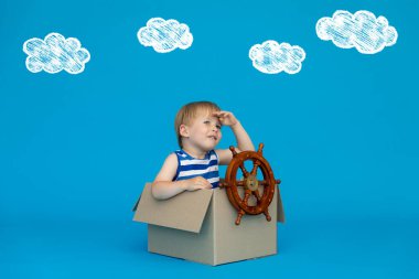 Mutlu çocuk kaptan olmayı hayal ediyor. Çocuk mavi arka planda eğleniyor. Çizgili tişörtlü çocuk karton kutuda oynuyor. Yaz tatili ve seyahat konsepti. Hayal ve hayal gücü