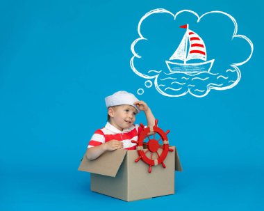 Mutlu çocuk kaptan olmayı hayal ediyor. Çocuk mavi arka planda eğleniyor. Çizgili tişörtlü çocuk karton kutuda oynuyor. Yaz tatili ve seyahat konsepti. Hayal ve hayal gücü