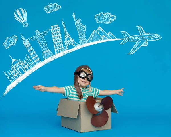 Happy child dreams of becoming a pilot. Kid having fun against blue paper background. Boy wearing striped shirt playing in cardboard box. Summer vacation and travel concept. Seven wonders of the world
