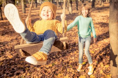 Mutlu çocuklar sonbahar parkında eğleniyorlar. Çocuklar arka planda sarı bulanık yapraklara karşı salıncakta oynuyorlar. Özgürlük ve tasasız konsept