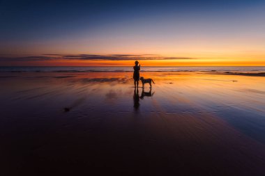 Bir fotoğraf yanında onu köpek ayakta ile sahilde canlı bir gün batımı sırasında çekici bir kadın silueti. Batı Avustralya, Avustralya.