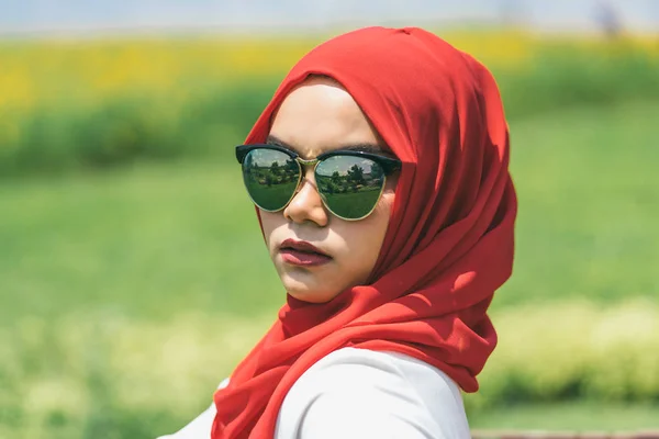 Retrato de la joven mujer musulmana feliz hiyab rojo sobre difuminado el campo verde con flores cosmos amarillas bajo el fondo azul del cielo . —  Fotos de Stock