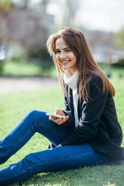 Doğada bir çim üzerinde oturan telefon ile gülümseyen güzel kadın