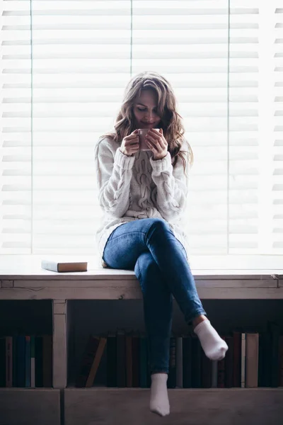 熱い飲み物を飲むと自宅の窓枠に座っている笑顔の女性 — ストック写真