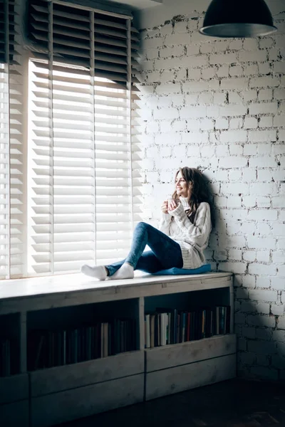 熱い飲み物を飲むと自宅の窓枠に座っている陽気な女性 — ストック写真