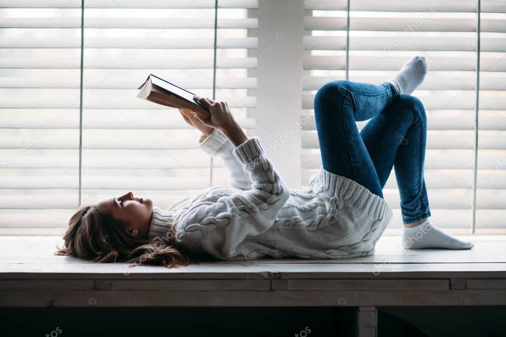  woman reading book lying on window sill at home