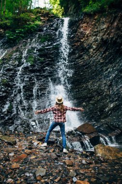 Şapkalı Kız kayanın üzerinde duruyor ve şelale tadını çıkarın. Pitoresk yere düşen su arasında genç bayan. Yaban hayatı arka plan arkadan genç kız portresi