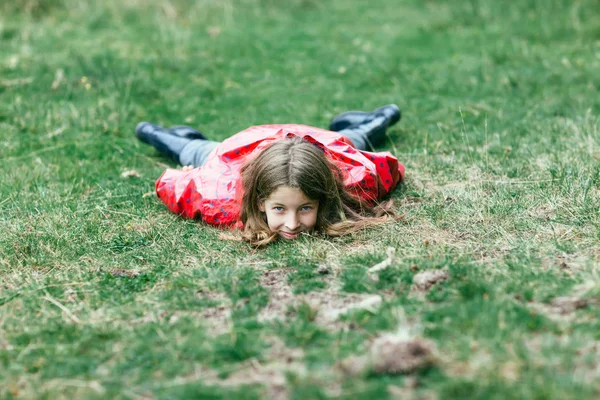 カメラに頭の芝生の上で横になっているピンクのジャケットの女の子 芝生の上の腹の上に横たわる 直視して笑顔の子 白人ヌード雨コートとゴムでは オープンエアのリラックス ブーツします — ストック写真