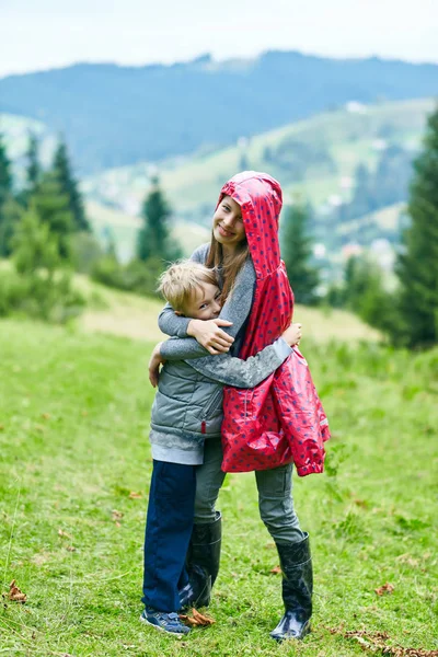 Brother hugging with sister in pink raincoat outdoors. Happy children standing on green meadow looking at camera on background of scenic landscape. Funny family members, boy and girl, on tourist walk.