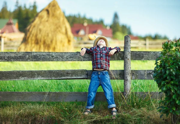 Boy leaned his back on wooden fence, head thrown back. Kid in farm clothes posing against a wooden fence and smiling. Portrait of young farmer looking at sky in summer sunny day