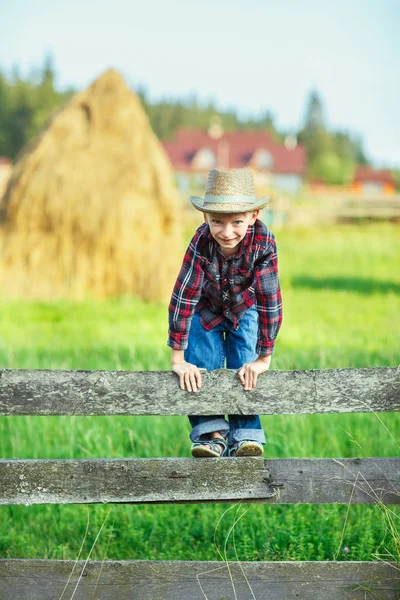 小男孩爬过木栅栏 直视着 闭上了 孩子在户外玩干草背景的木制篱笆 勇敢的小牛仔摆出一个危险的姿势 — 图库照片
