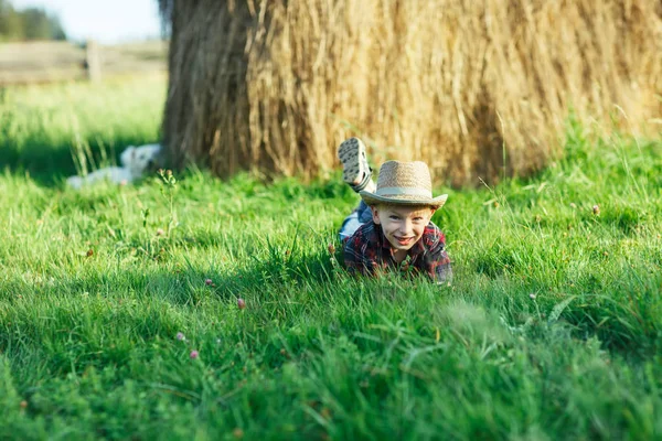 ハンサムな男の子は干し草の背景の草にあります 子供は自然にかかっている 手で彼の胃と Propps の頭の上にあります 若い男が屋外の肖像画 週末の概念 — ストック写真