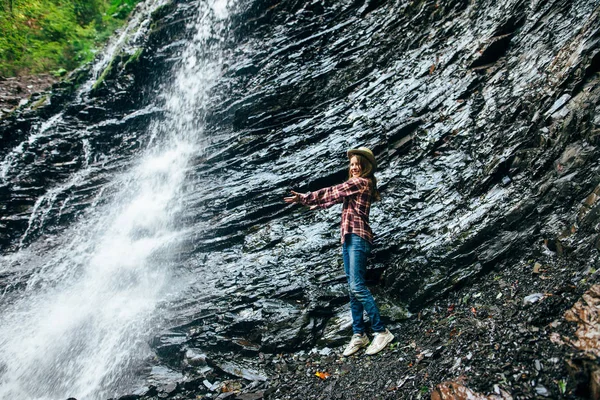 年轻的女孩站在瀑布附近 伸出双臂拥抱它 十几岁的年轻人在水中摆姿势 在镜头前微笑 休息在狂放的自然 徒步旅行 夏天概念 — 图库照片