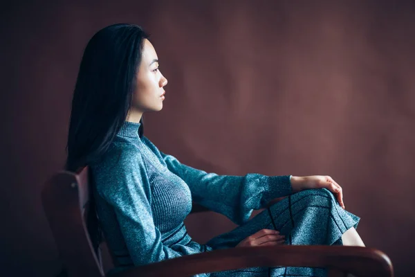 Profile portrait of asian beautiful woman sitting in chair. Eastern model in blue dress on brown background