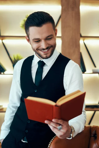 Przystojny Uśmiechnięty Młodzieniec Eleganckim Garniturze Czytający Książkę Nowoczesnym Luksusowym Wnętrzu — Zdjęcie stockowe