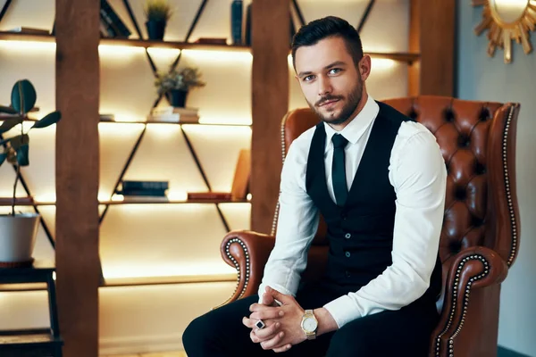 Handsome young man sitting and posing in armchair in modern luxury interior