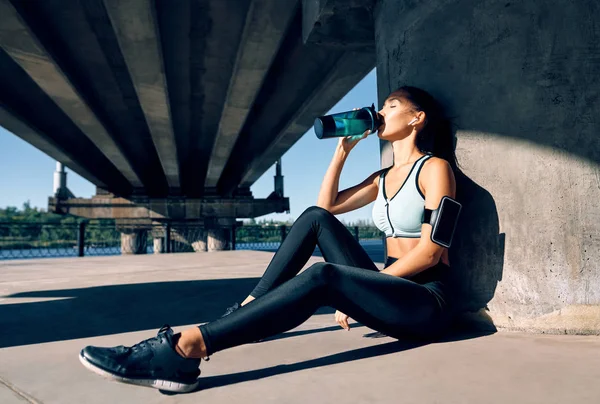 年轻健康的女人从瓶子里喝水 在靠近河流的工业城市背景下锻炼后的体态苗条的女性休息 — 图库照片