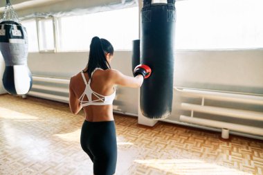 Bayan boksörün fitness salonundaki kum torbasına vuruşunun arka görüntüsü. Kadın boks stüdyosunda yumruk pratiği yapıyor.                  