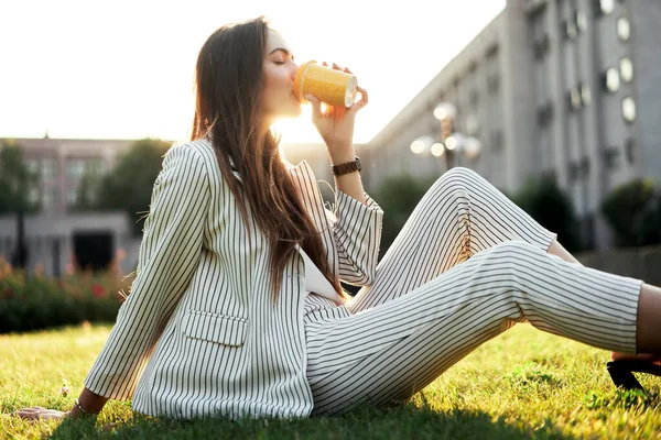 年轻时尚的女人坐在城市公园的草地上 沐浴在阳光下 一边喝着长颈鹿 一边放松 休息时间概念 — 图库照片