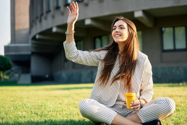 Young pretty woman welcome friends waving raised palm, sitting on grass with coffee. Hello,   greeting concept