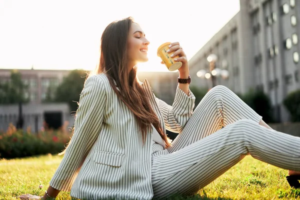 若いスタイリッシュな女性はコーヒーを飲みながら 日差しの中で都市公園の芝生の上に座ってリラックスしてください 休憩時間の概念 — ストック写真