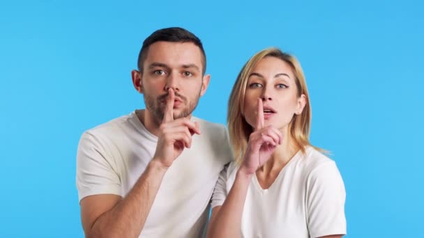 一对年轻夫妇把手指放在嘴唇上 做着沉默的手势 把阴谋隔离在蓝色的背景下 沉默的概念 — 图库视频影像