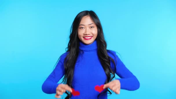 Щаслива молода жінка тримає два маленьких паперових серця очима ізольовані на синьому фоні — стокове відео