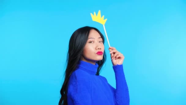 Vacker asiatisk kvinna med papper krona på pinne poserar på blå bakgrund — Stockvideo