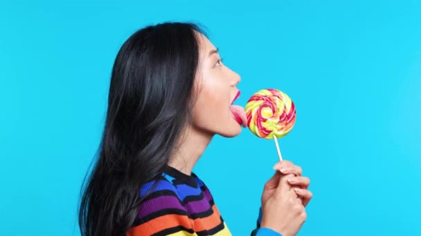 Profile view of young woman licking colorful lollipop on blue background — Stock Video