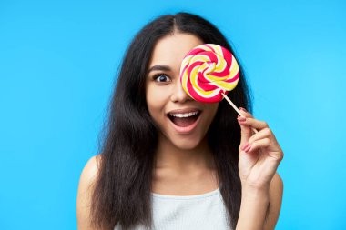 Heyecanlı mutlu siyah kadın mavi arka planda izole edilmiş renkli lolipopla gözlerini kapatıyor. Duygular, eğlence, yemek konsepti