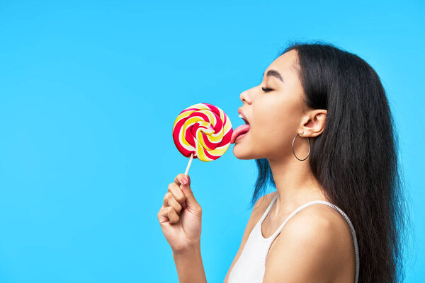 Профиль вид молодой черной женщины облизывающей красочный леденец на синем фоне с копировальным пространством. Еда, концепция развлечений
