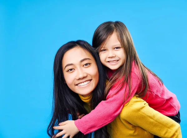 Mãe feliz e pequena filha piggybacking juntos isolados no fundo azul . — Fotografia de Stock