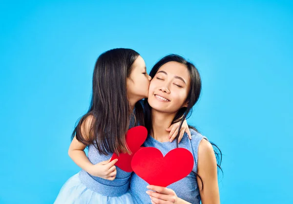 Gadis kecil mencium ibunya memegang hati kertas di latar belakang biru — Stok Foto