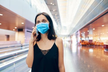 Korunan yüz maskeli kadın modern alışveriş merkezinde ya da havaalanında akıllı telefondan konuşuyor.