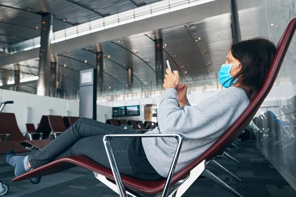 Vrouw in virus bescherming gezichtsmasker rusten met smartphone in de luchthaven van vertrek lounge — Stockfoto