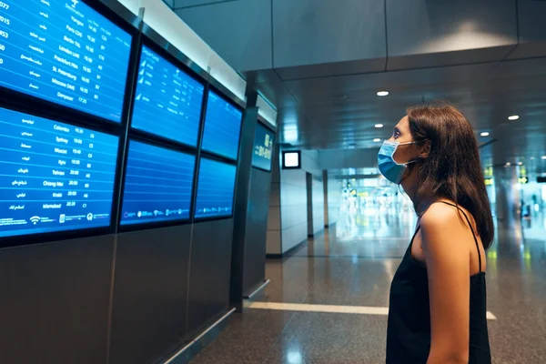 Mulher em máscara de proteção de vírus olhando para a placa de informações verificando seu voo no aeroporto internacional — Fotografia de Stock
