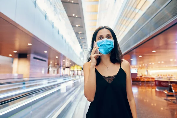 Woman in protection face mask speaking on smartphone in modern shopping mall or airport