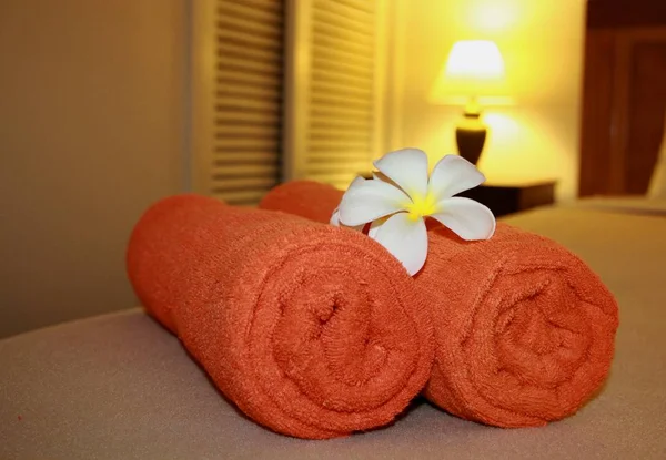 Two bright orange towels rolled and placed on the bed in a hotel spa, with a white plumeria flower on top
